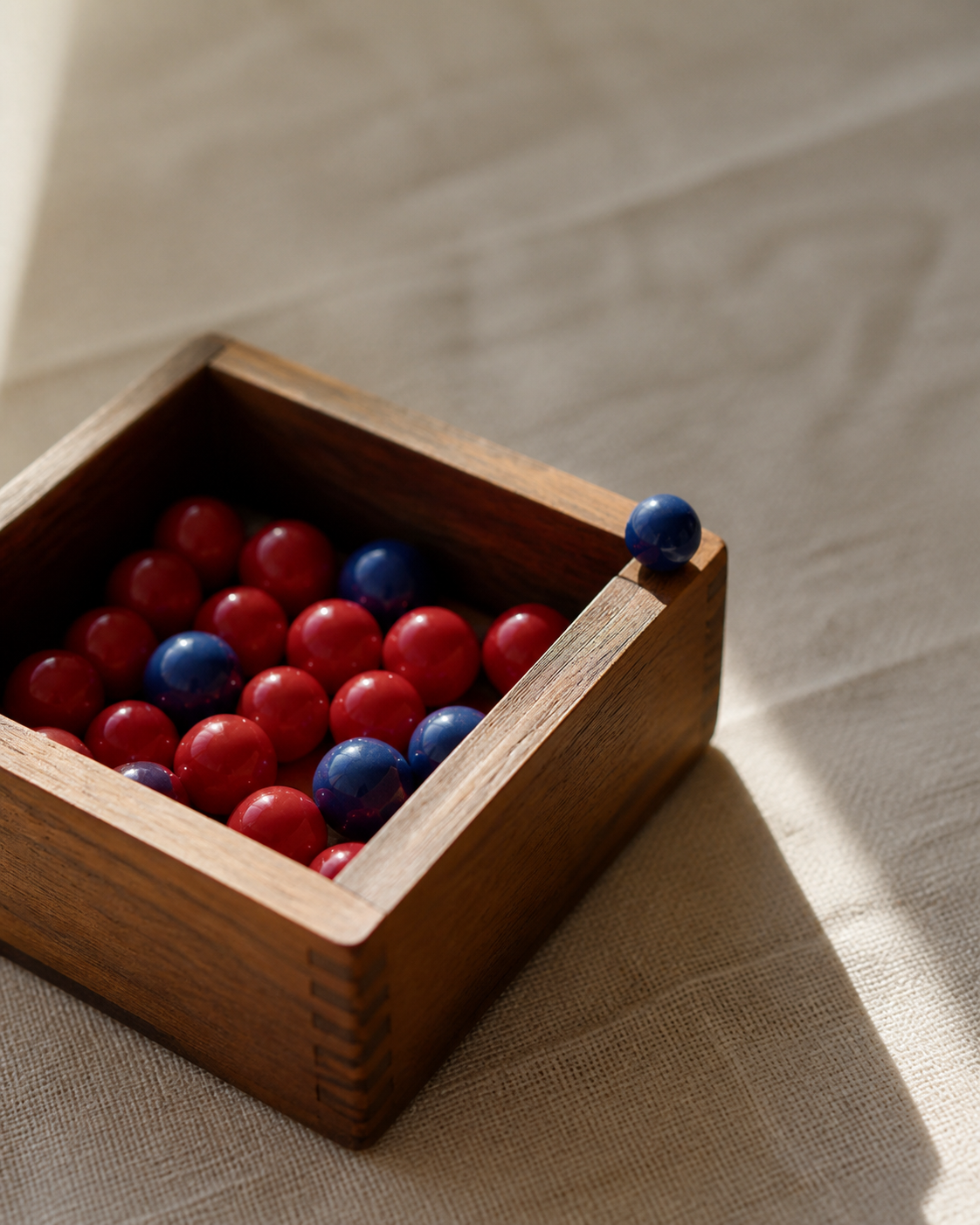A polished wooden box of marbles split into red and blue, with one blue marble pulled out and resting on the rim, lit like a still life.
