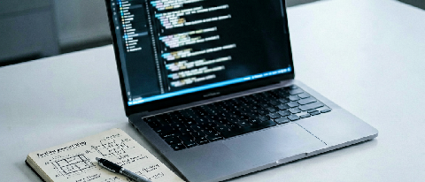 A minimalist desk setup featuring a laptop displaying abstract code patterns and a physical notebook with handwritten technical diagrams, symbolizing the transi