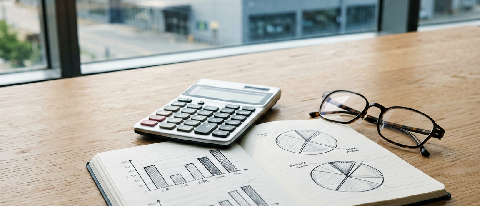 A minimalist desk setup featuring a calculator, a notebook with hand-drawn cost charts, and a blurred background of a modern tech building.