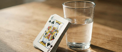 A stack of playing cards leaning against a glass of water on a wooden table, warm editorial lighting
