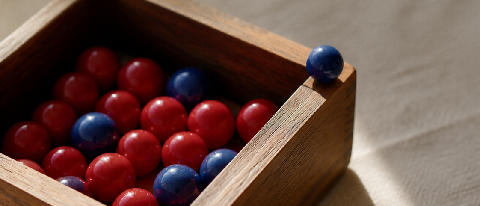 A polished wooden box of marbles split into red and blue, with one blue marble pulled out and resting on the rim, lit like a still life.
