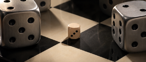 A small wooden die with Arabic numeral '3' sitting on a giant gaveled chessboard, surrounded by much larger silver dice.