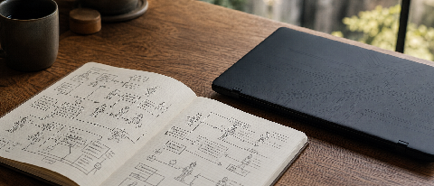 A minimalist desk setup showing a notebook with hand-drawn architectural diagrams next to a closed laptop, symbolizing human strategic thinking over automated c