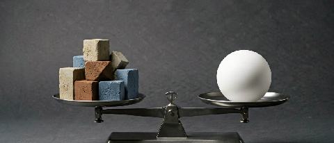 Editorial still life showing a precision balance scale with multiple distinct geometric weights on one side and a single large weight on the other, symbolizing 