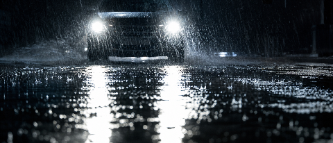 A car driving at night on a rain-soaked road with deep puddles reflecting headlights, shot from a low angle.